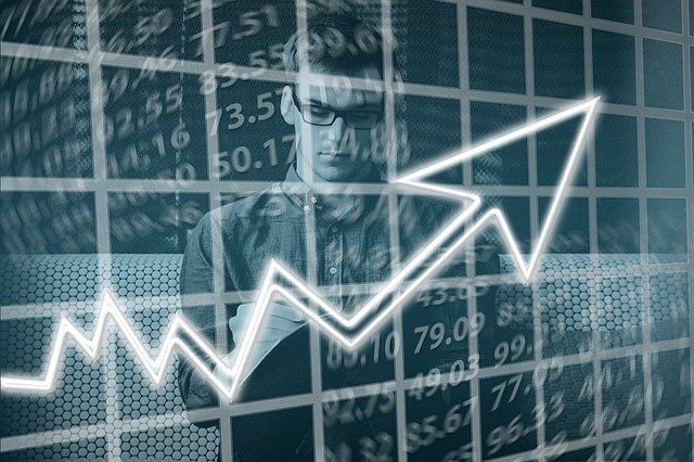 Photo of a young man, shwn from the waist up. He's wearing and buttoned shirt and glasses. In front of him, there is a graphic of an arrow on a graph with jagged lines. The arrow is climbing up toward the right, signifying growth or improvement.